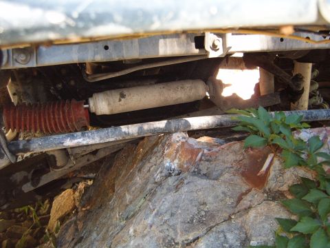 1988 Ford Bronco - Close-up view of the undercarriage, focusing on a shock absorber and its red bellows, with rocks and foliage visible.