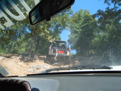 1988 Ford Bronco - Two vehicles navigate a challenging off-road trail surrounded by trees under a clear blue sky.
