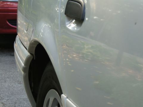 1998 Ford Taurus - The rear quarter panel and wheel of a silver 1998 Ford Taurus are visible, with another red vehicle in the background.
