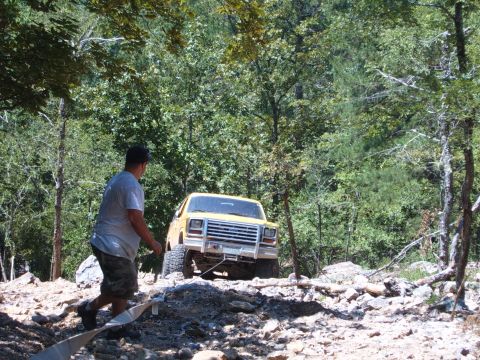 1988 Ford Bronco - A yellow SUV navigates a rocky, wooded trail with a person assisting.