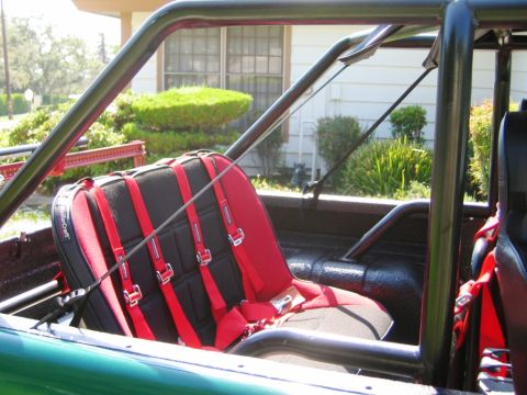 1985 Ford Bronco - Vehicle interior shows racing seats and a roll cage, with a house and greenery in the background.
