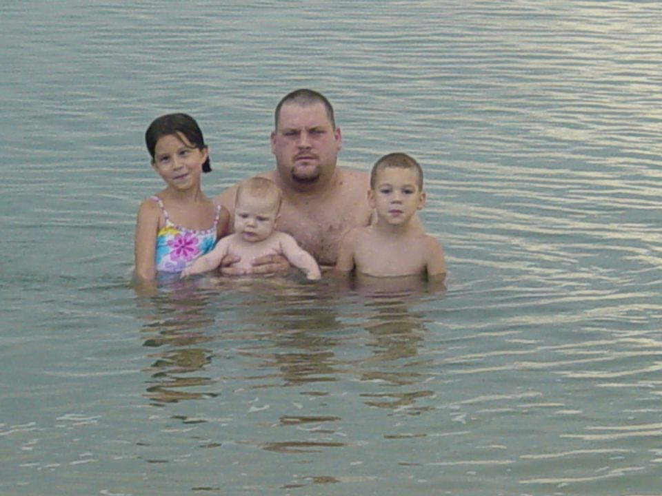 Family of four in shallow water, man holding a baby, with two children beside him
