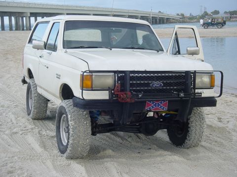 1989 Ford Bronco - A white 1989 Ford Bronco with off-road tires parked on a sandy beach, with another vehicle visible in the background.