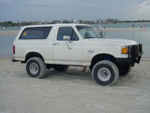 1989 Ford Bronco - White 1989 Ford Bronco sits on a sandy beach near the water with people in the distance.
