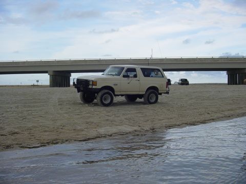 1989 Ford Bronco - A classic Ford Bronco sits on the sand near the water, with a bridge overhead and another vehicle in the distance.