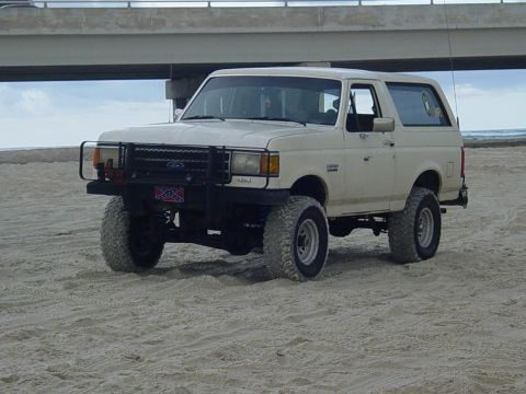 1989 Ford Bronco - This white Ford Bronco SUV is parked on a sandy beach, modified with a bull bar and off-road tires.