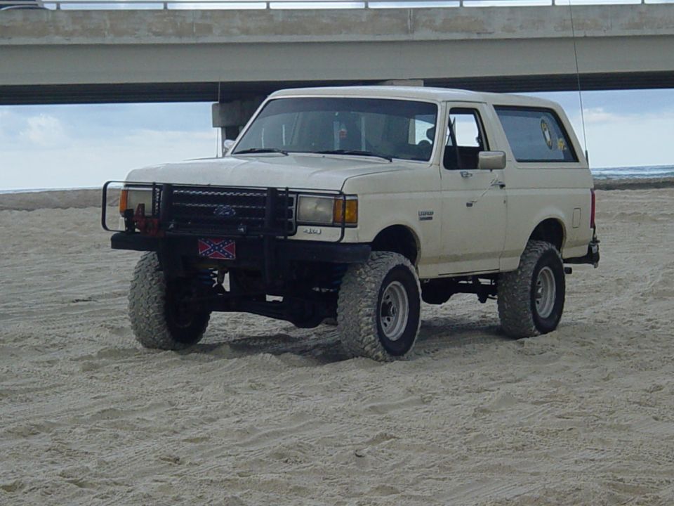 White Ford Bronco SUV with a black bull bar and off-road tires on a sandy beach
