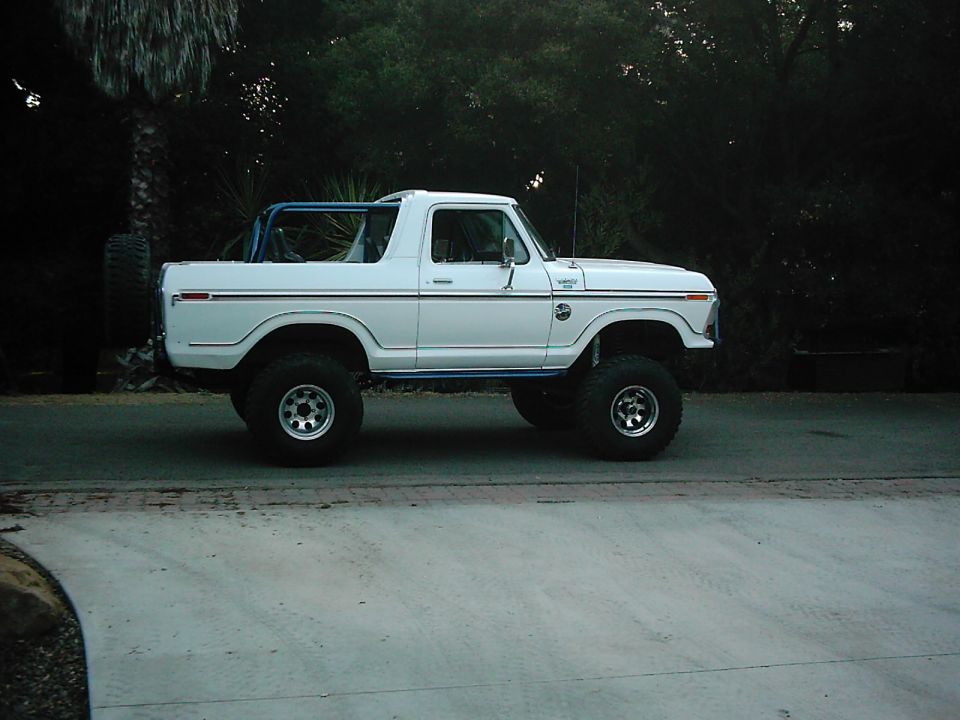 White 1978 Ford Bronco lifted with large tires and a blue roll cage, viewed from the side