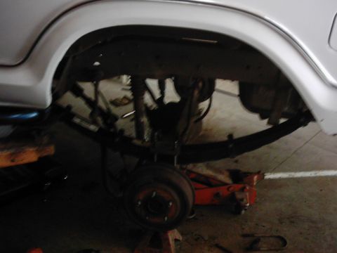 1978 Ford Bronco - The rear suspension of a white vehicle is shown, with a hydraulic jack supporting the axle and a brake drum visible.