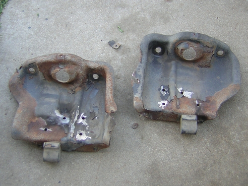 Two rusted metal undercarriage parts with holes drilled in them, lying on concrete