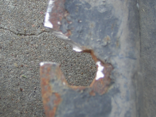 Close-up of a dark, rusted metal part with a hexagonal cutout against a cracked asphalt background