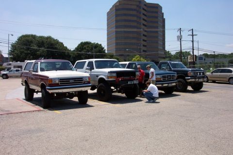1992 Ford Bronco - Four lifted full-size Ford Broncos parked nose-out in a row at a club meet in a paved lot, spanning maroon-over-white, white, blue-over-sil…