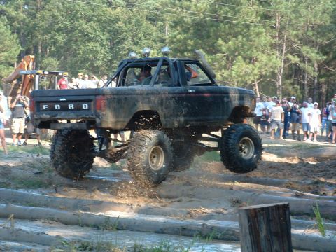 1993 Ford Bronco - This modified Ford Bronco is airborne over a muddy obstacle course, with spectators gathered to watch the event.
