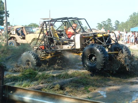 1993 Ford Bronco - A custom off-road buggy navigates a muddy track, kicking up spray as another vehicle watches from the background.