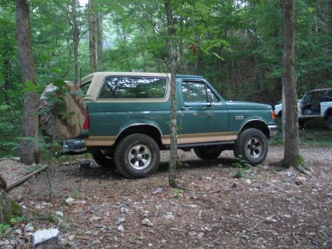 1995 Ford Bronco - A green Ford Bronco is parked off-road in a wooded area, with another vehicle partially visible in the background.