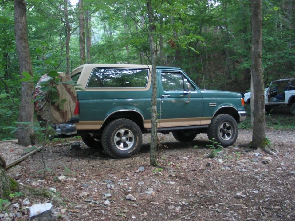 Green Ford Bronco parked in woods, spare tire on back. Another white SUV is visible behind it