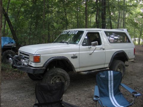 1995 Ford Bronco - A white Ford Bronco is parked in a wooded area with camping chairs nearby and another vehicle in the background.