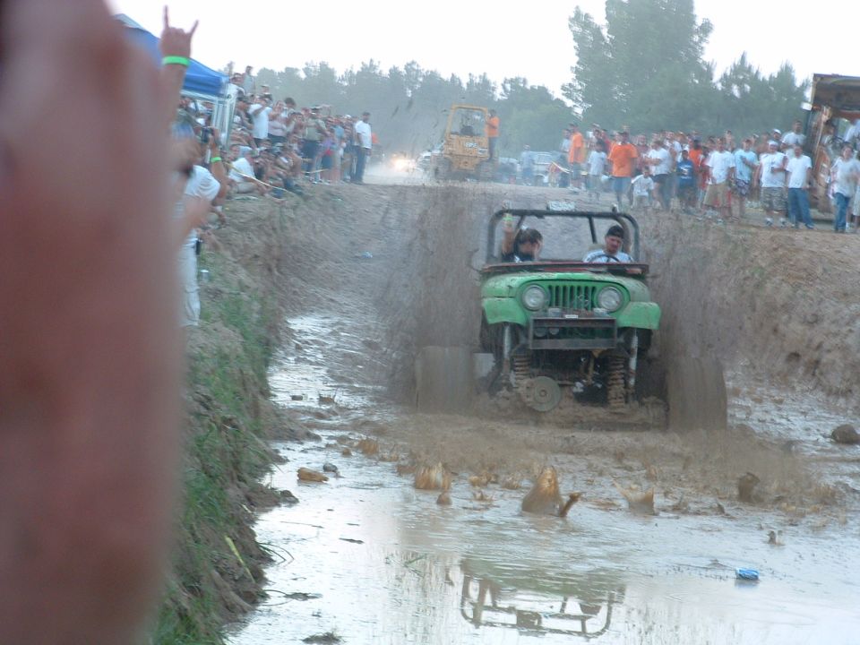 Green jeep driving through mud pit with spectators on the bank