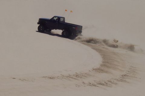 1994 Ford F-150 - A pickup truck drives on a sandy dune, creating a trail of kicked-up sand.