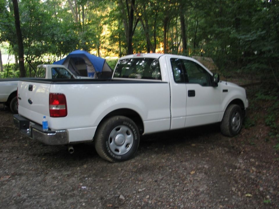 white pickup truck parked next to a blue tent in a wooded area, another white vehicle is partially visible to the left