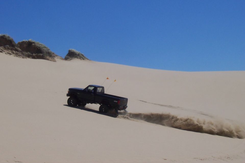 Black pickup truck driving on a sand dune, kicking up dust. The truck has a lifted suspension and off-road tires