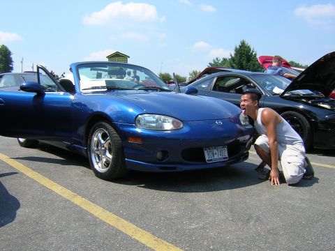 1995 Ford Probe - A man playfully interacts with a blue Mazda Miata convertible, parked next to another car.