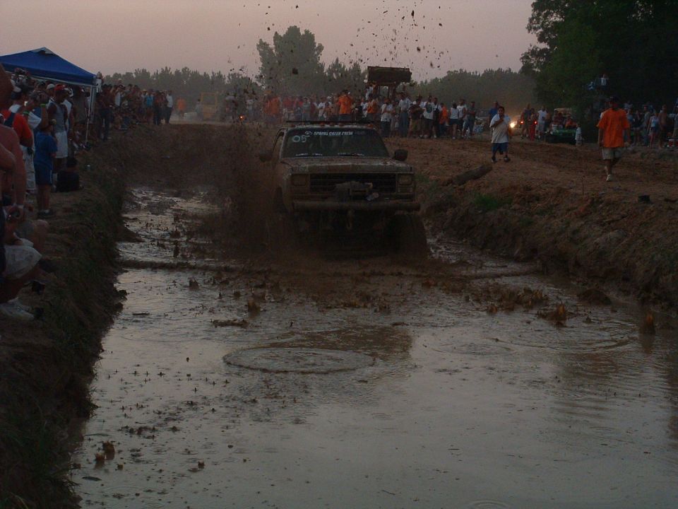 Mud-splattered 1980s Ford Bronco driving through a muddy track with spectators watching