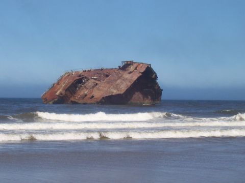 1994 Ford F-150 - A rusted shipwreck rests on the shore, battered by ocean waves under a clear blue sky.