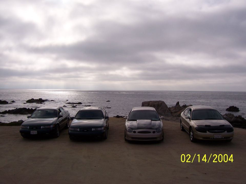 Four sedans parked on a dirt lot overlooking the ocean, one is a 1998 Mercury Sable