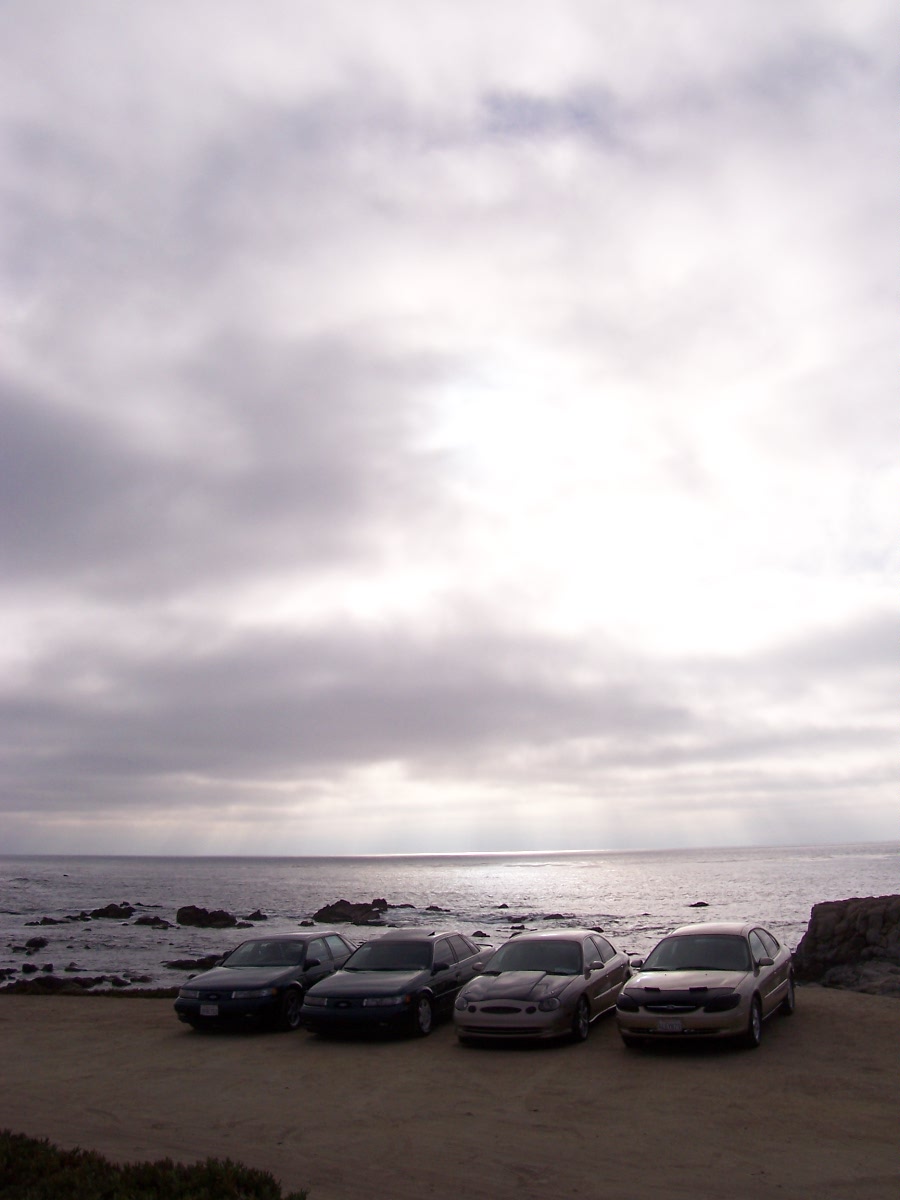four sedans parked on dirt lot facing ocean, cloudy sky
