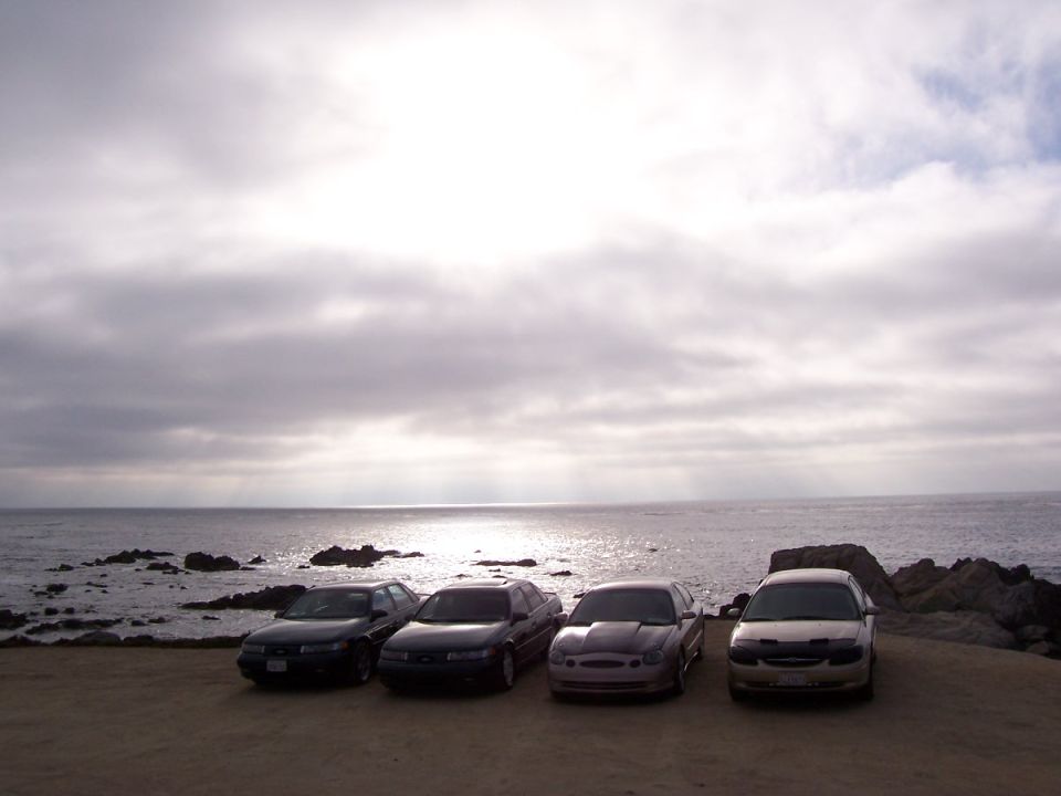 Four sedans parked on a dirt lot by the ocean, with a cloudy sky