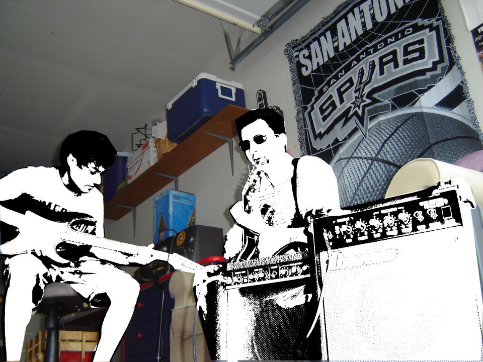 Two guys are playing guitars in a garage. One is sitting and playing a guitar. The other is standing and playing a guitar next to an amplifier