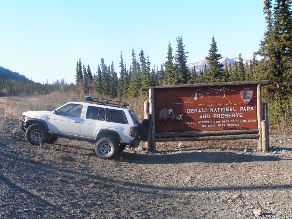 White lifted Nissan Pathfinder beside the Denali National Park entrance sign, side view