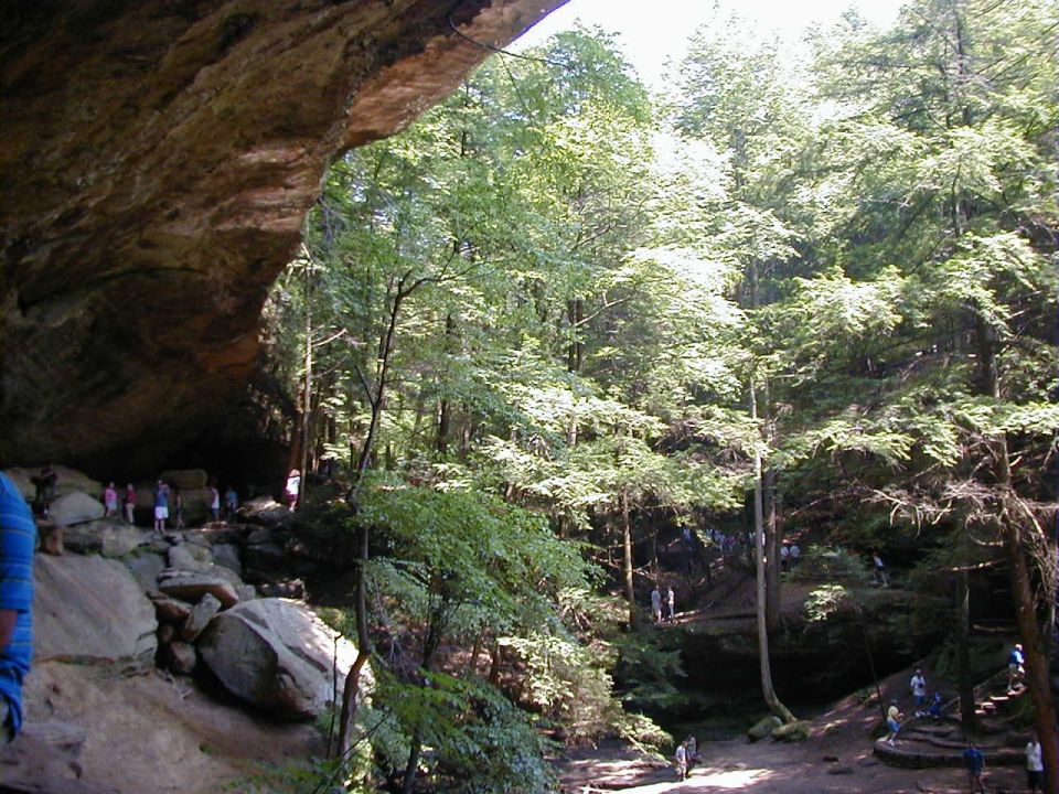 Forest scene with a large rock overhang and a stream, people are visible on the rocks and trails