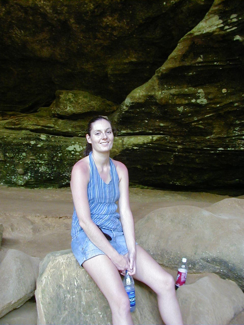 Woman in blue striped top and denim shorts sits on rocks inside a cave, holding a water bottle