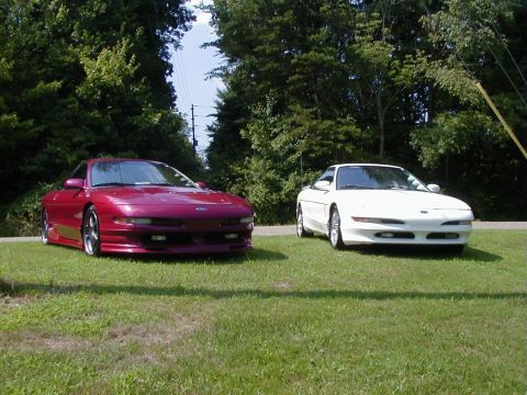 1994 Ford Probe - Two Ford Probe cars, one maroon and one white, are parked on a grassy area with trees in the background.