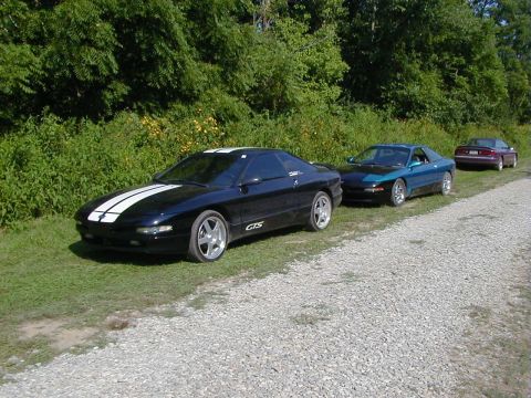 1994 Ford Probe - Three Ford Probes and a sedan are parked in a line on a gravel road next to a grassy area.