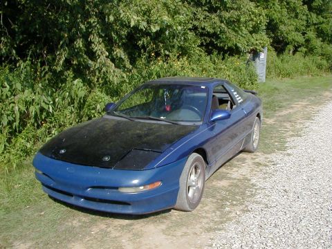 1994 Ford Probe - A blue coupe with a black hood sits on a gravel road, surrounded by greenery.