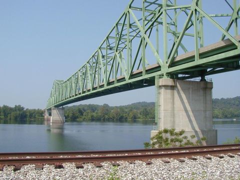 1994 Ford Probe - A large green truss bridge spans a calm river, with railroad tracks running alongside the water.