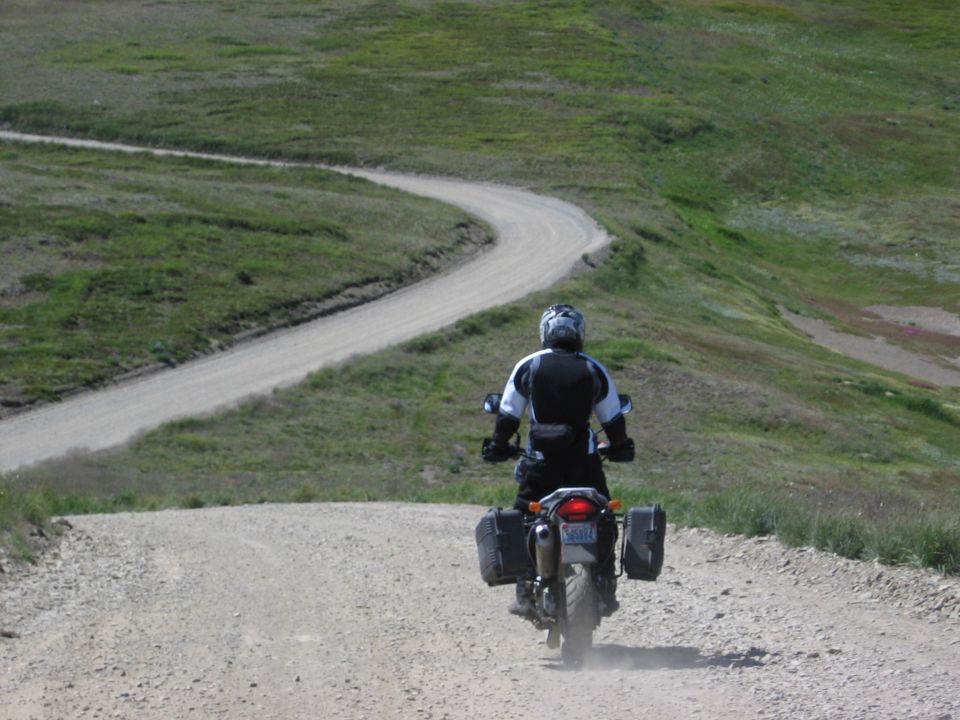 Rear view of black adventure motorcycle with panniers on winding gravel road through green hills