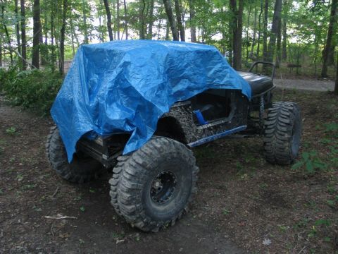 1995 Ford Bronco - A modified off-road vehicle sits partially covered by a blue tarp in a forest setting.