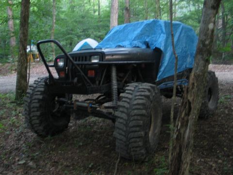 1995 Ford Bronco - A heavily modified Jeep Wrangler sits in a forest clearing, partially covered by a blue tarp.