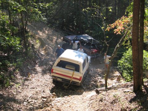 1995 Ford Bronco - Two off-road vehicles navigate a challenging rocky trail in a wooded area.