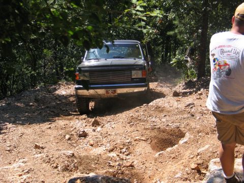 1995 Ford Bronco - A dark SUV navigates a challenging off-road path surrounded by trees.