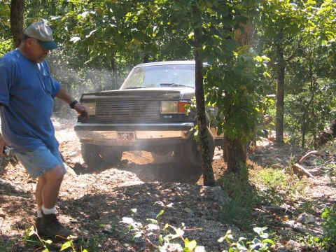 1995 Ford Bronco - A man stands by a Ford Bronco as it drives through a wooded area on a dirt path.
