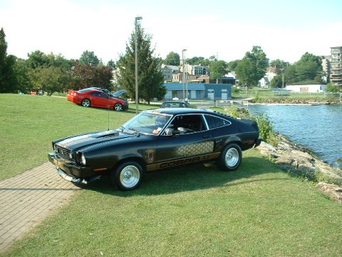 1978 Ford Mustang II - A black 1978 Ford Mustang II is parked on a grassy area next to a body of water, with another red car in the background.