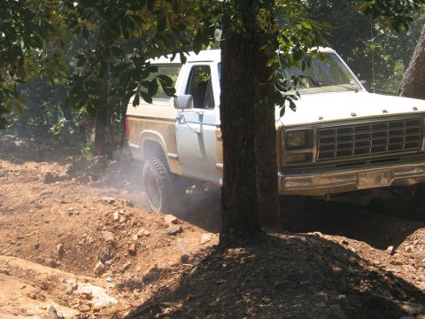 1995 Ford Bronco - Ford Bronco navigating an off-road trail with trees and dirt mounds.