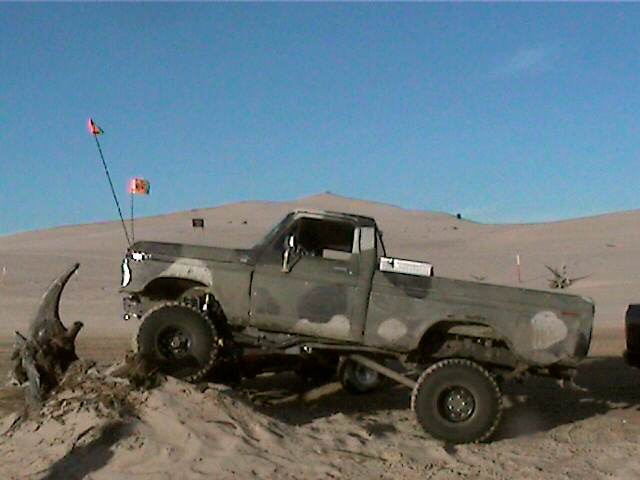 Muddy camo lifted Ford pickup with whip flag stuck in sand dunes