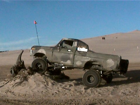 1977 Ford F-150 - A lifted pickup truck with camouflage paint is shown on a sand dune under a clear blue sky.
