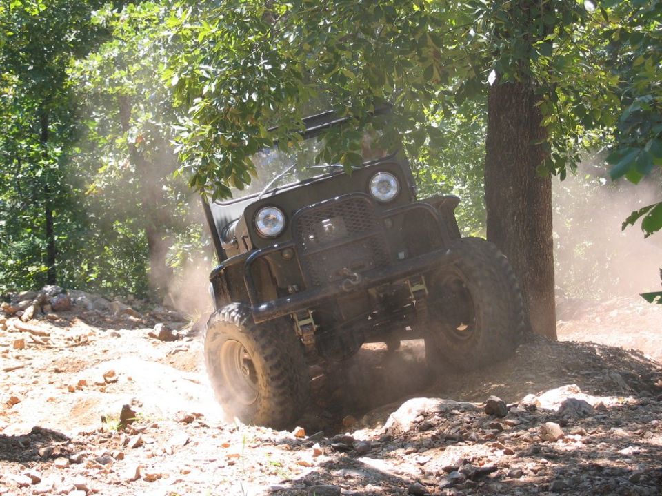 Dark green off-road vehicle with large tires navigating a dusty, rocky trail under trees